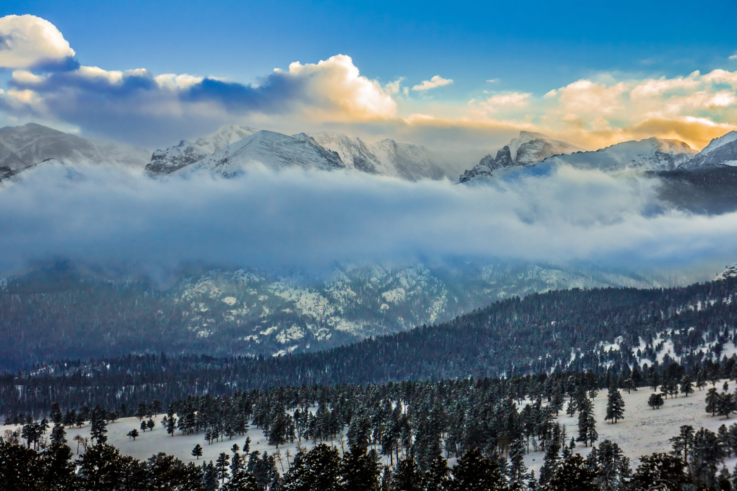 Snow-covered mountains rise above a forested landscape with low clouds partially obscuring the peaks under a blue sky with scattered clouds.