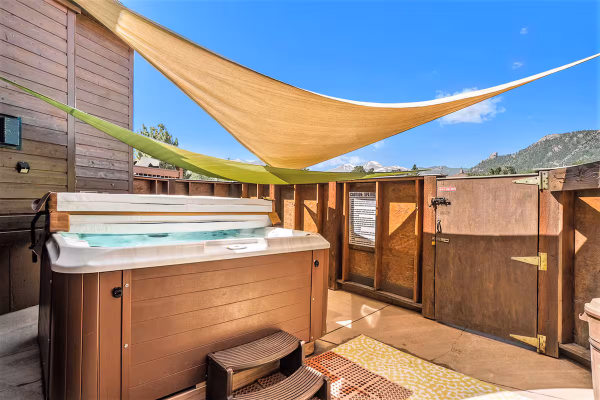 A hot tub sits on a concrete patio, surrounded by wooden privacy walls. Shade sails are stretched overhead, and a small rug is placed near the tub. Mountains and blue sky are visible in the background.