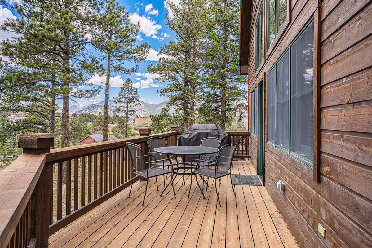 A wooden deck attached to a cabin features a round table with four metal chairs and a covered grill, surrounded by tall pine trees and a view of distant mountains under a partly cloudy sky.
